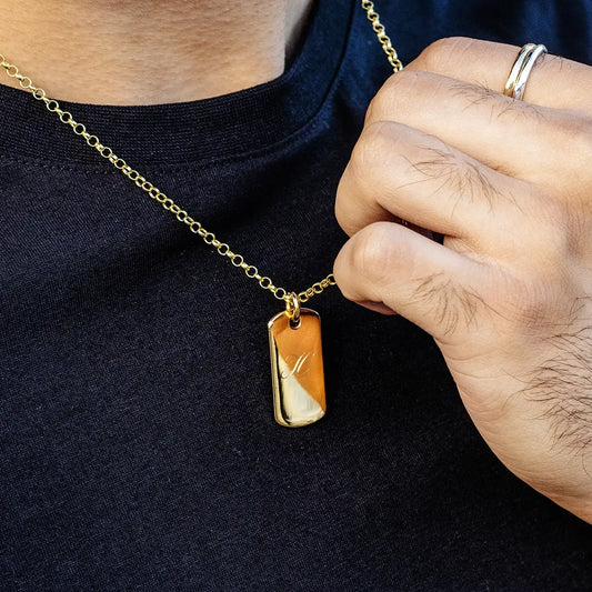 Close-up of a person holding a gold dog tag pendant necklace on a belcher chain, engraved with an initial, worn over a dark top