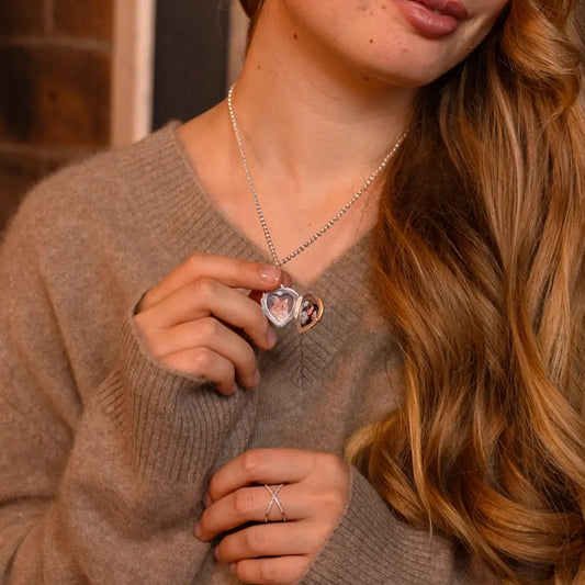 Close-up of a woman wearing a beige sweater, holding open a silver heart locket necklace that reveals two small photo inserts.