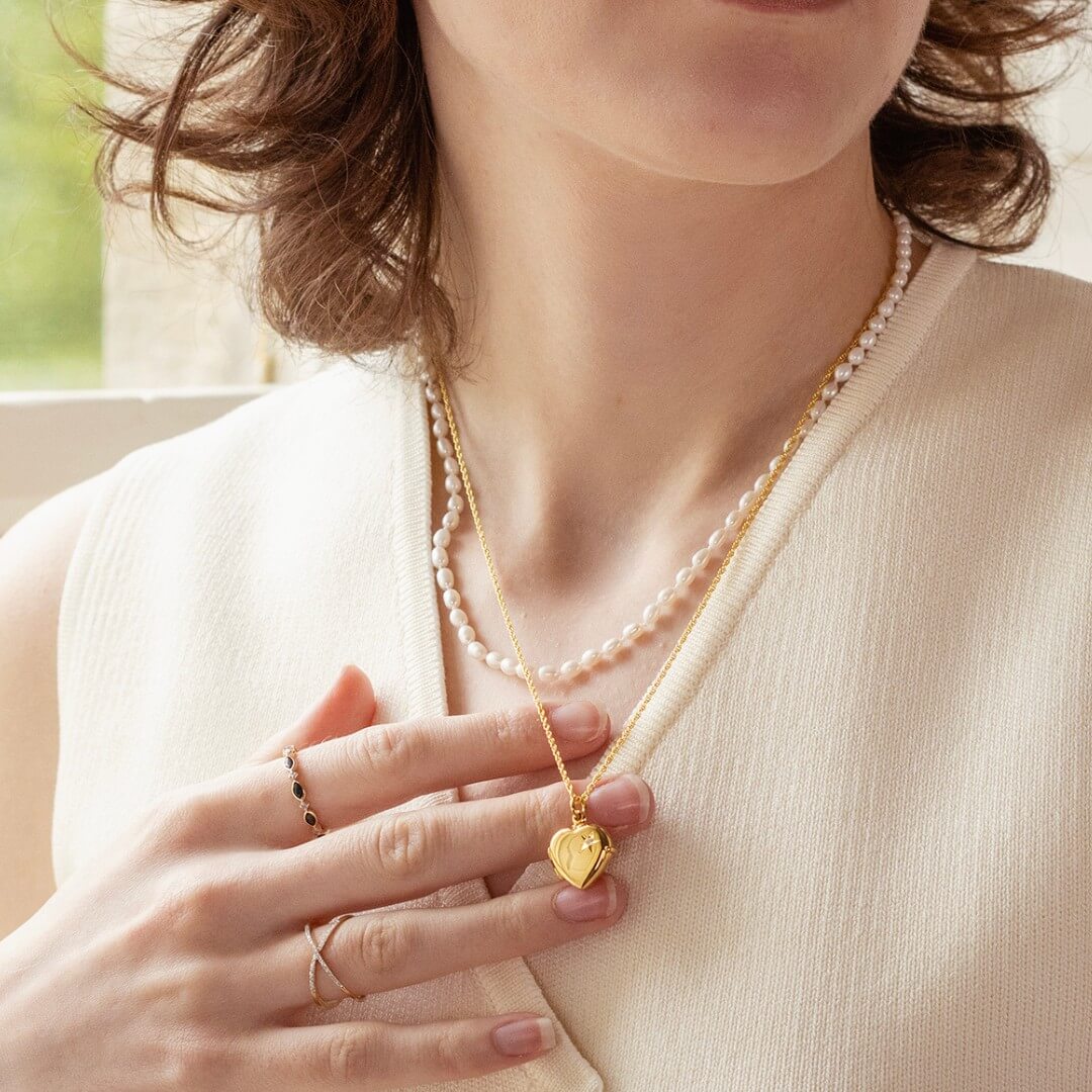 Woman wearing a 2 photo diamond heart locket in gold and a pearl necklace, holding the gold locket