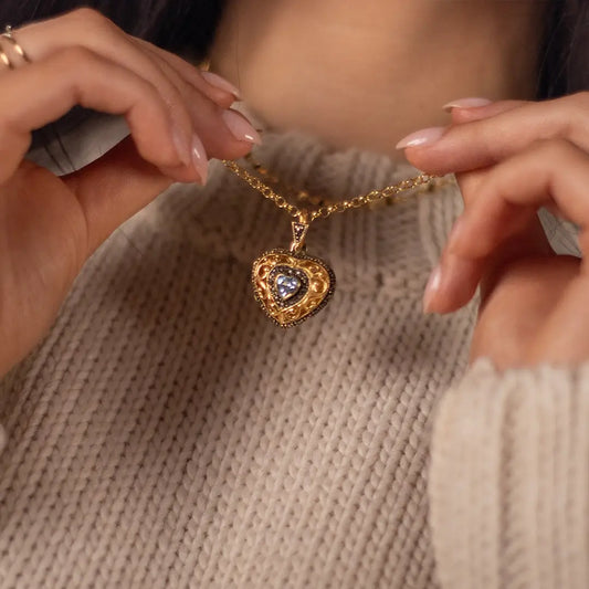 A person holding a gold heart-shaped locket with ornate filigree design and blue topaz gemstone centre, against a cream knit sweater.