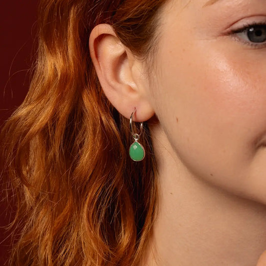 A close-up of a person with red hair wearing a silver hoop earring with a chrysoprase teardrop stone against a dark reddish-brown background.