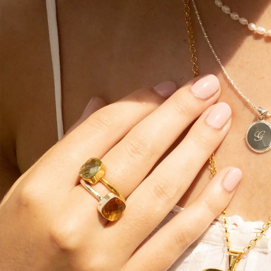 Close-up of gold and silver gemstone ring with green and amber stones, styled with engraved initial pendant necklace