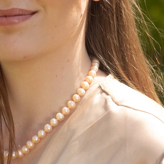 Close-up of a woman wearing a champagne pearl necklace, featuring softly lustrous round pearls resting along the neckline against a neutral blouse in natural sunlight.