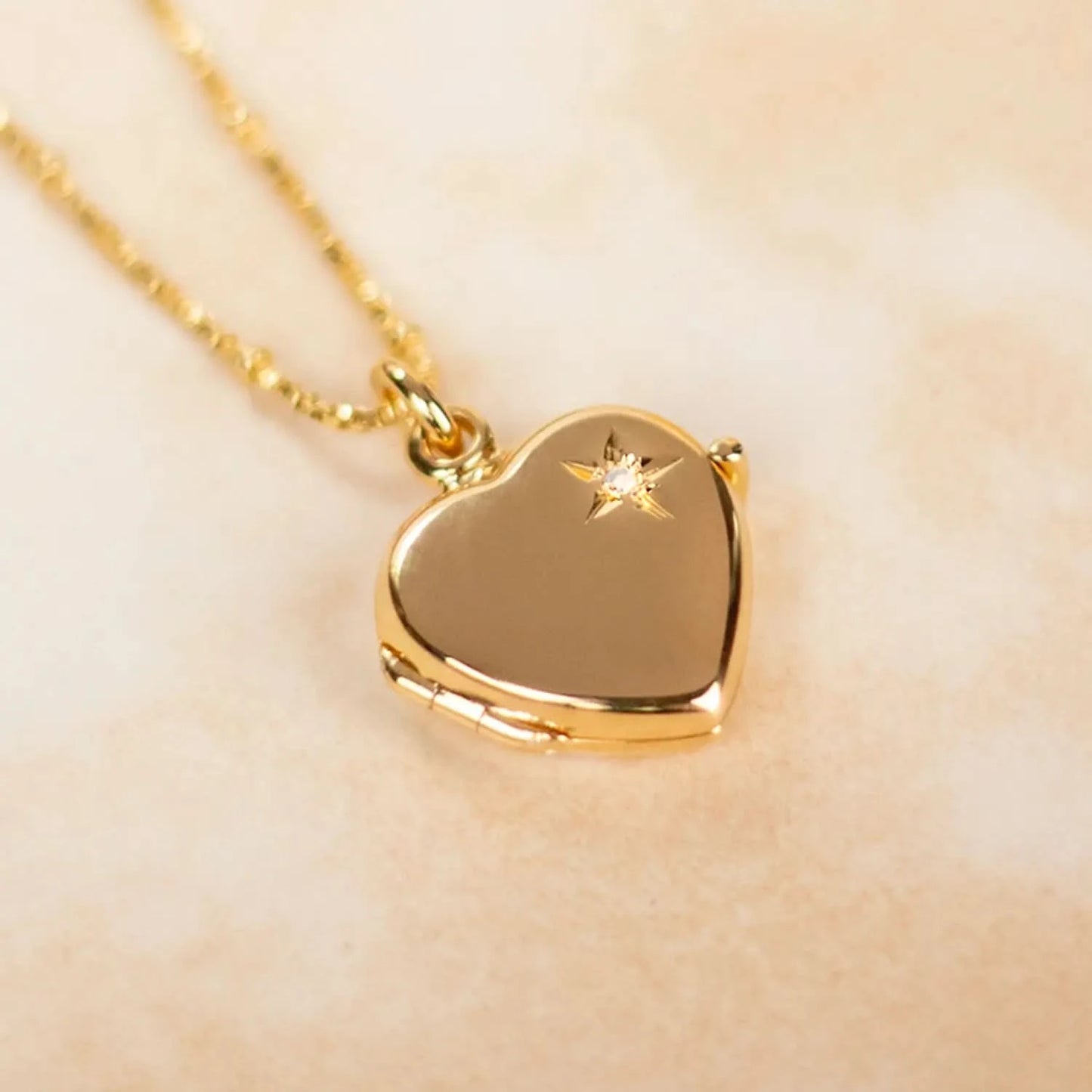 lifestyle image of a gold heart-shaped locket necklace featuring a delicate starburst engraving with a central diamond. Shown against a soft, neutral background
