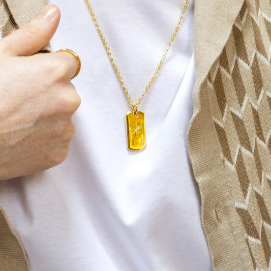 Gold dog tag necklace with a yellow pendant held by a male hand against a white t-shirt