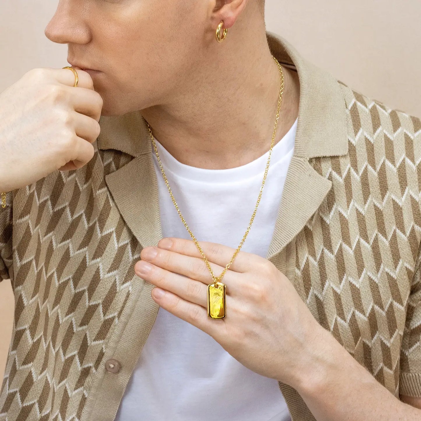 Man wearing a gold dog tag necklace with a yellow pendant, holding the necklace with their hand.