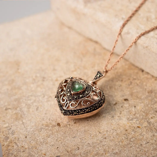 Close-up of a rose gold vintage-style heart locket necklace with an emerald centre stone and ornate detailing, displayed on stone.