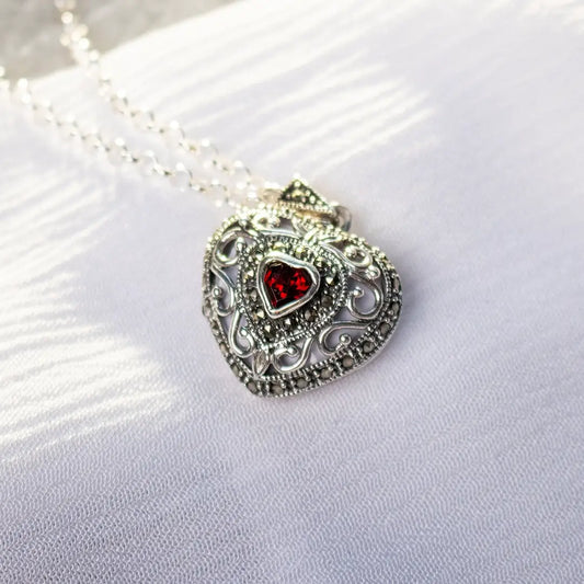 Sterling silver vintage heart locket necklace with ornate detailing and a red garnet gemstone, photographed on a soft white fabric background.