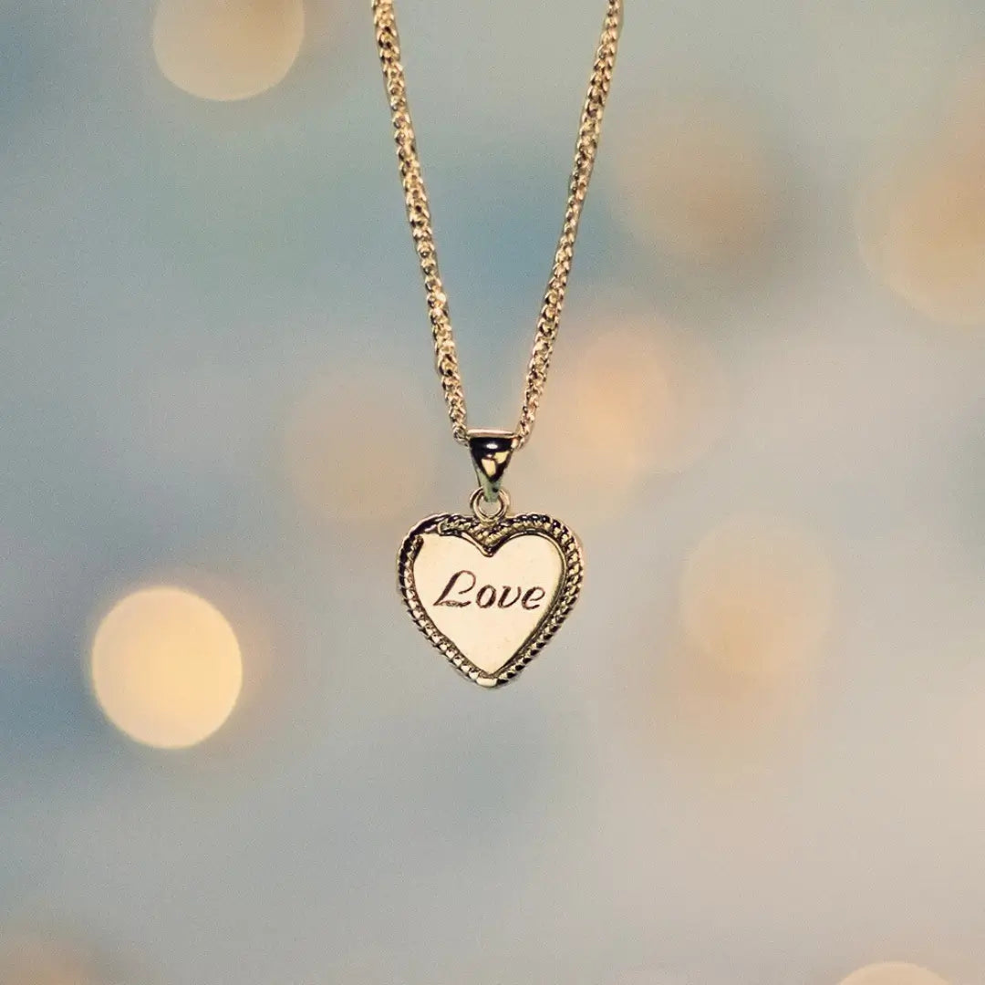 Gold heart-shaped pendant necklace engraved with the word ‘Love,’ hanging from a delicate chain against a soft, blurred background with warm light bokeh