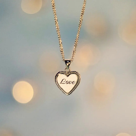 Gold heart-shaped pendant necklace engraved with the word ‘Love,’ hanging from a delicate chain against a soft, blurred background with warm light bokeh