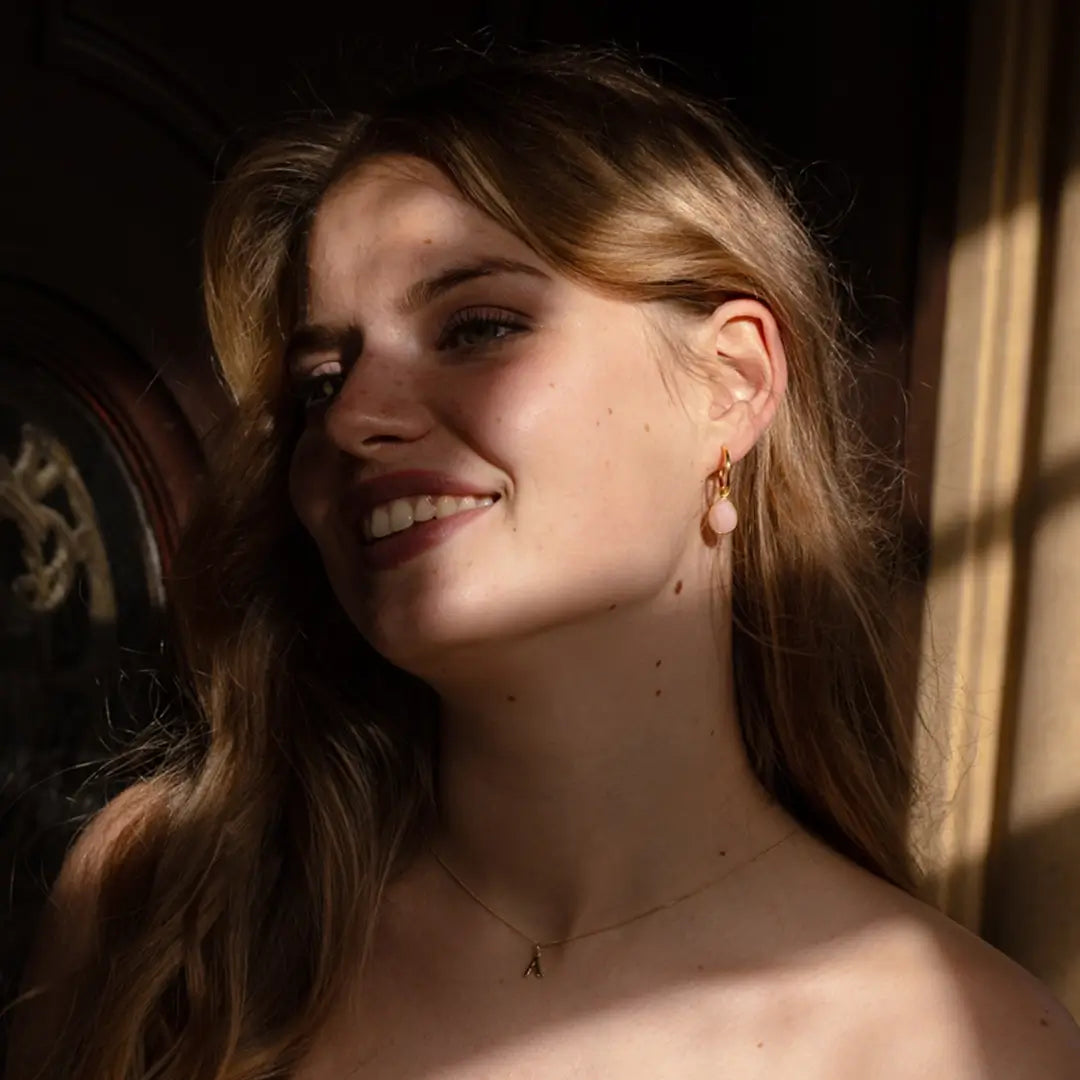 Woman smiling in soft sunlight, wearing gold hoop earrings with teardrop-shaped pink opal gemstones and a delicate gold initial necklace