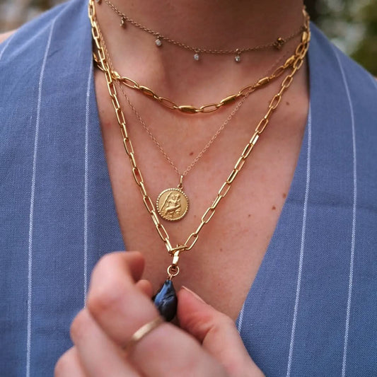 Lifestyle photo of a woman wearing layered gold necklaces, including a delicate St. Christopher medallion pendant. Styled with a mix of paperclip chains and a blue pinstripe outfit