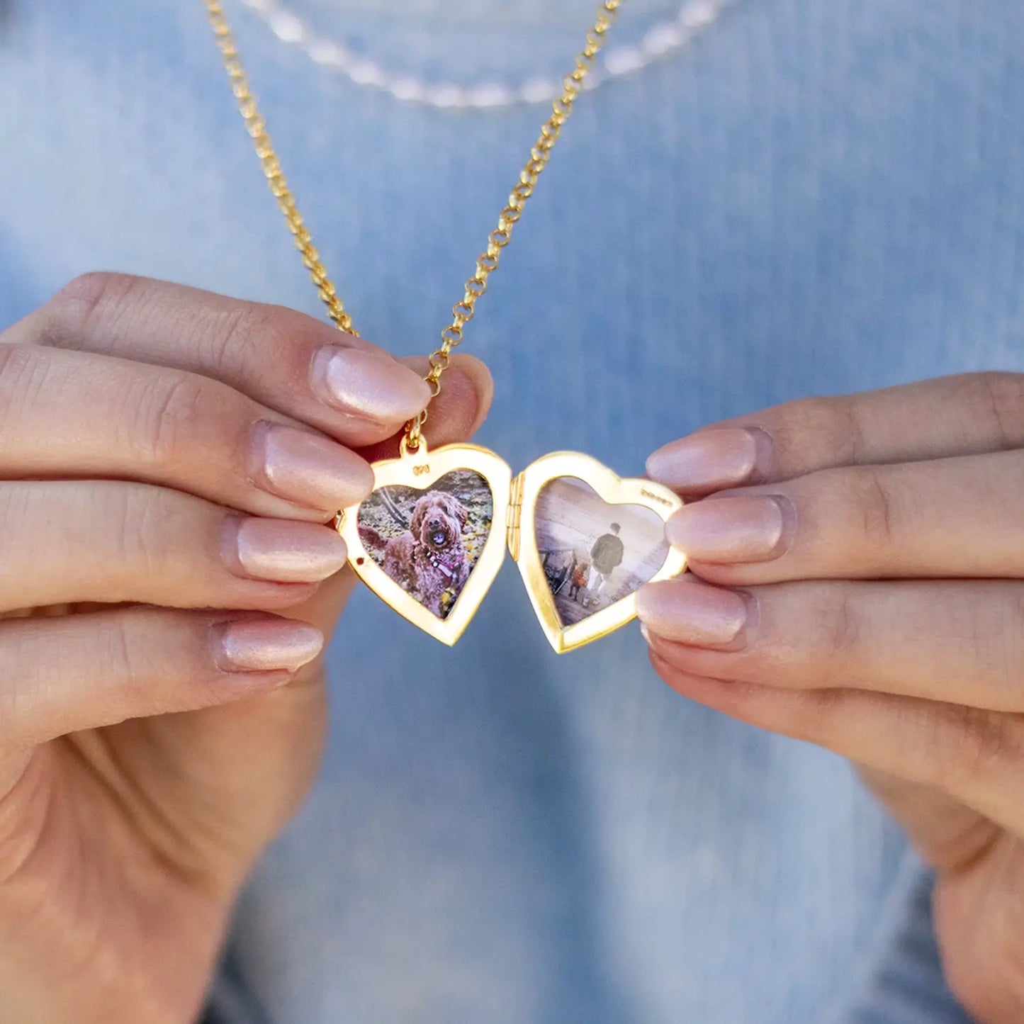 A close-up image of an open gold heart-shaped locket held between two hands, displaying two custom photo inserts: one of a dog and one of a person on a beach. The locket hangs from a polished belcher chain, and the wearer is dressed in a soft blue top