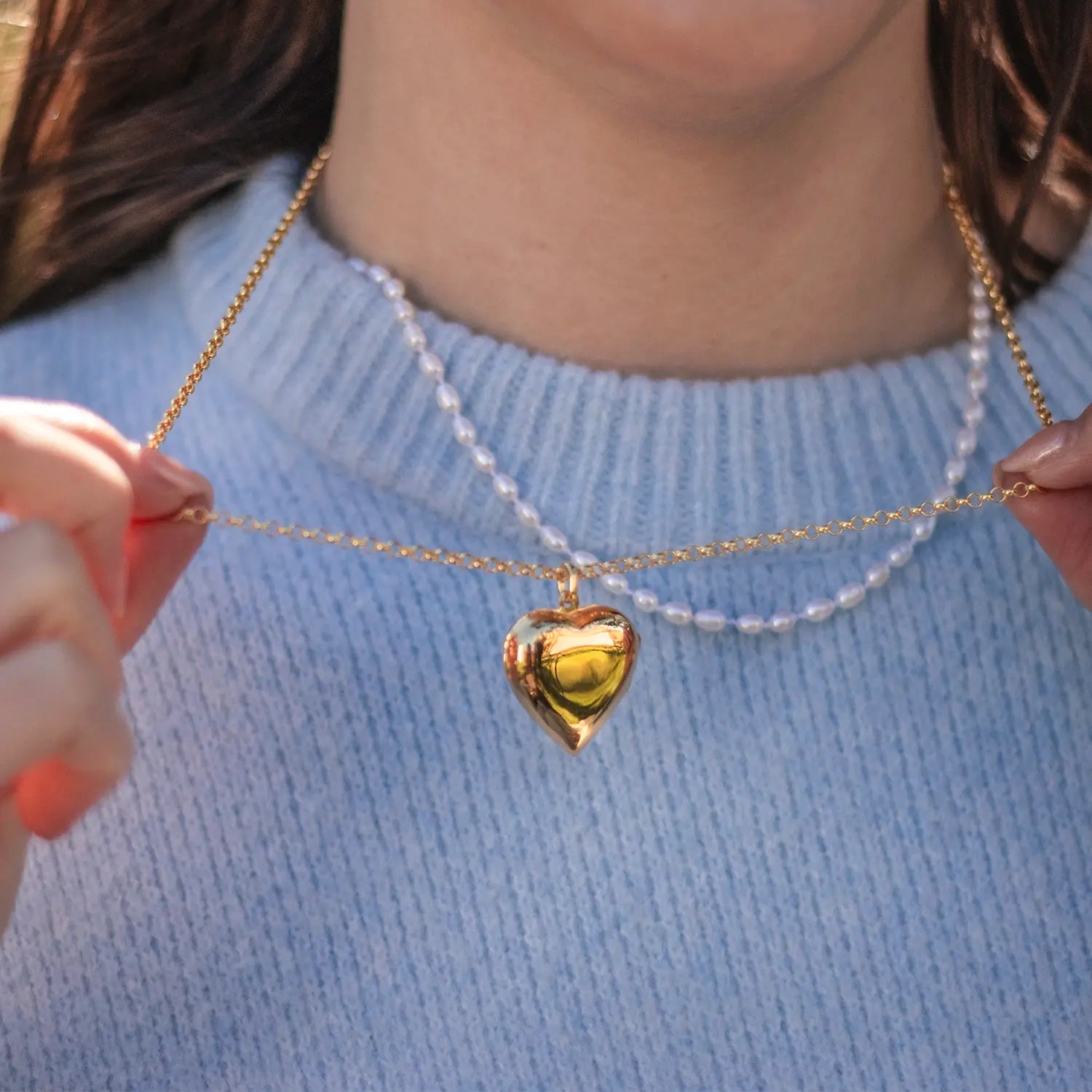 Model holding gold heart locket necklace on belcher chain over light blue sweater
