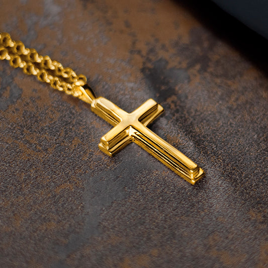 Gold cross pendant necklace on a chain, photographed close-up on a textured dark surface, highlighting the polished finish and clean, classic design.