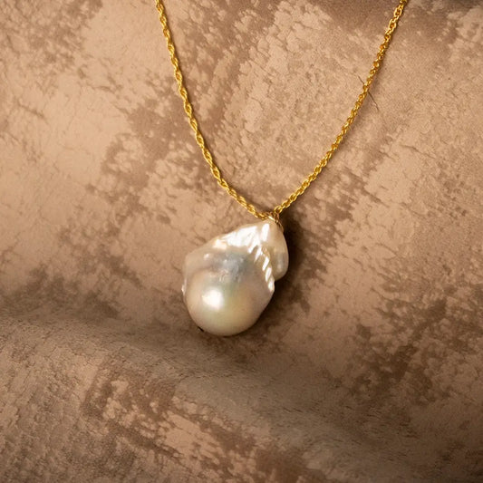 Ivory baroque pearl pendant necklace on a gold chain, photographed on textured neutral fabric background.