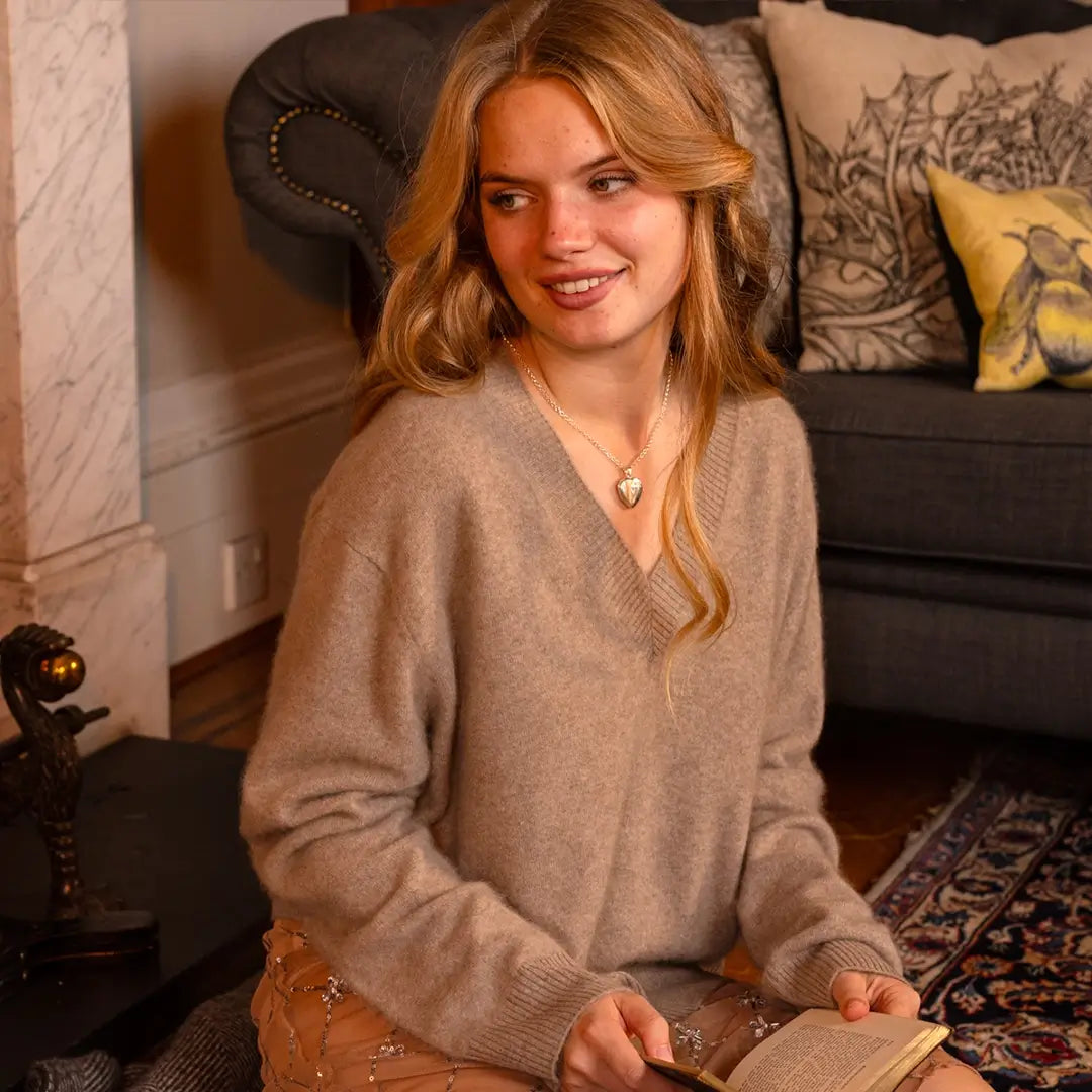 A woman wearing a beige sweater and soft waves in her hair smiles while holding a book, accessorized with a silver heart locket necklace.