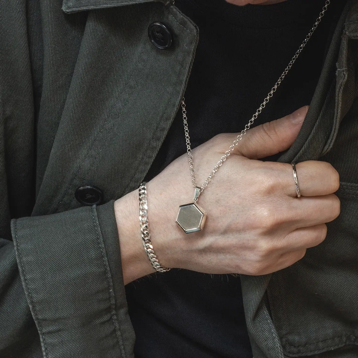 Close-up of man wearing a sterling silver Cuban link bracelet, hexagon pendant necklace, and silver ring with a dark jacket.