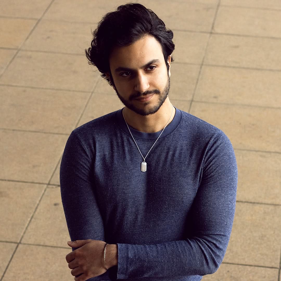 Man wearing silver dog tag necklace and blue long-sleeve shirt, arms crossed