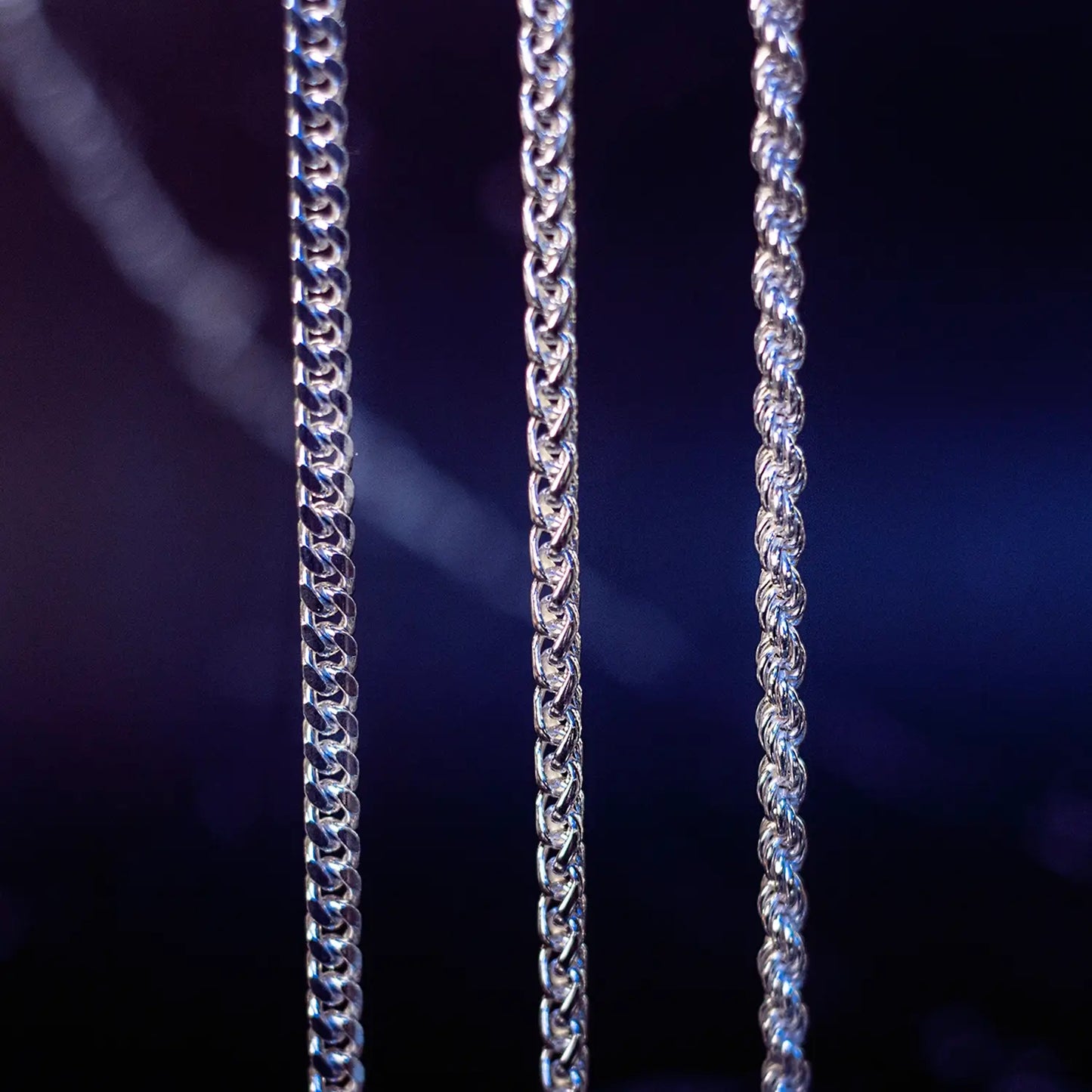 Close-up of three sterling silver chain designs showing different link styles under blue lighting.