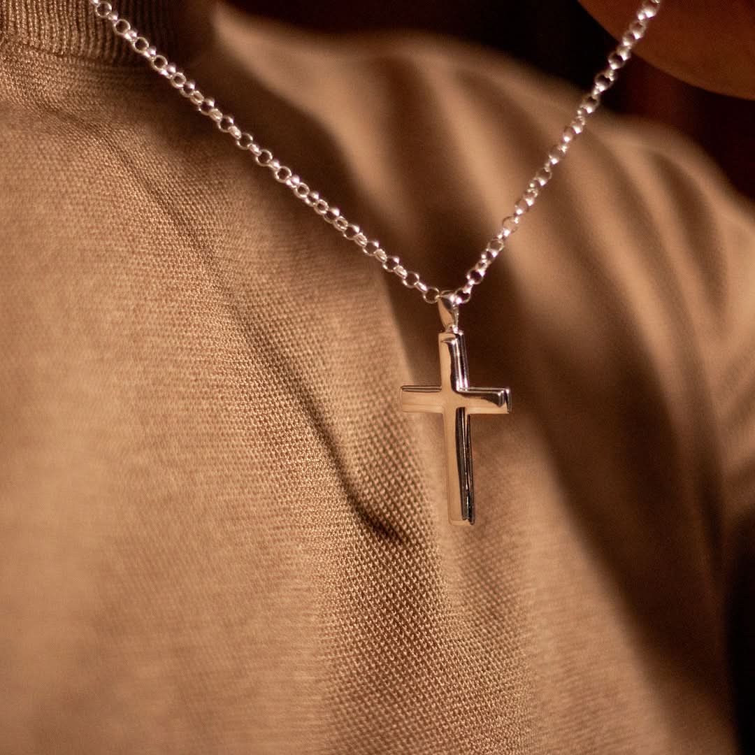 close up photograph of a heavy weight silver cross necklace on a belcher chain, worn by a male model wearing a brown jumper