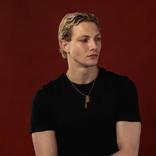 Male model wearing a gold double dog tag necklace against a dark red background