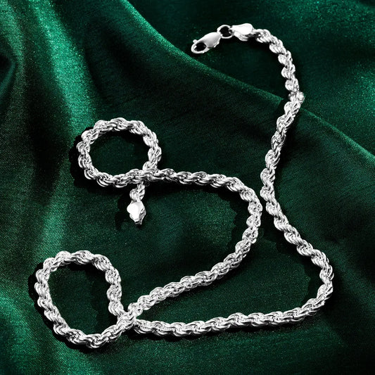 A silver rope-style chain and matching bracelet displayed on a deep green satin fabric. The twisted links create a textured, reflective surface, and both pieces have lobster-claw clasps. Soft highlights and shadows emphasize the draped fabric and the shine of the metal.