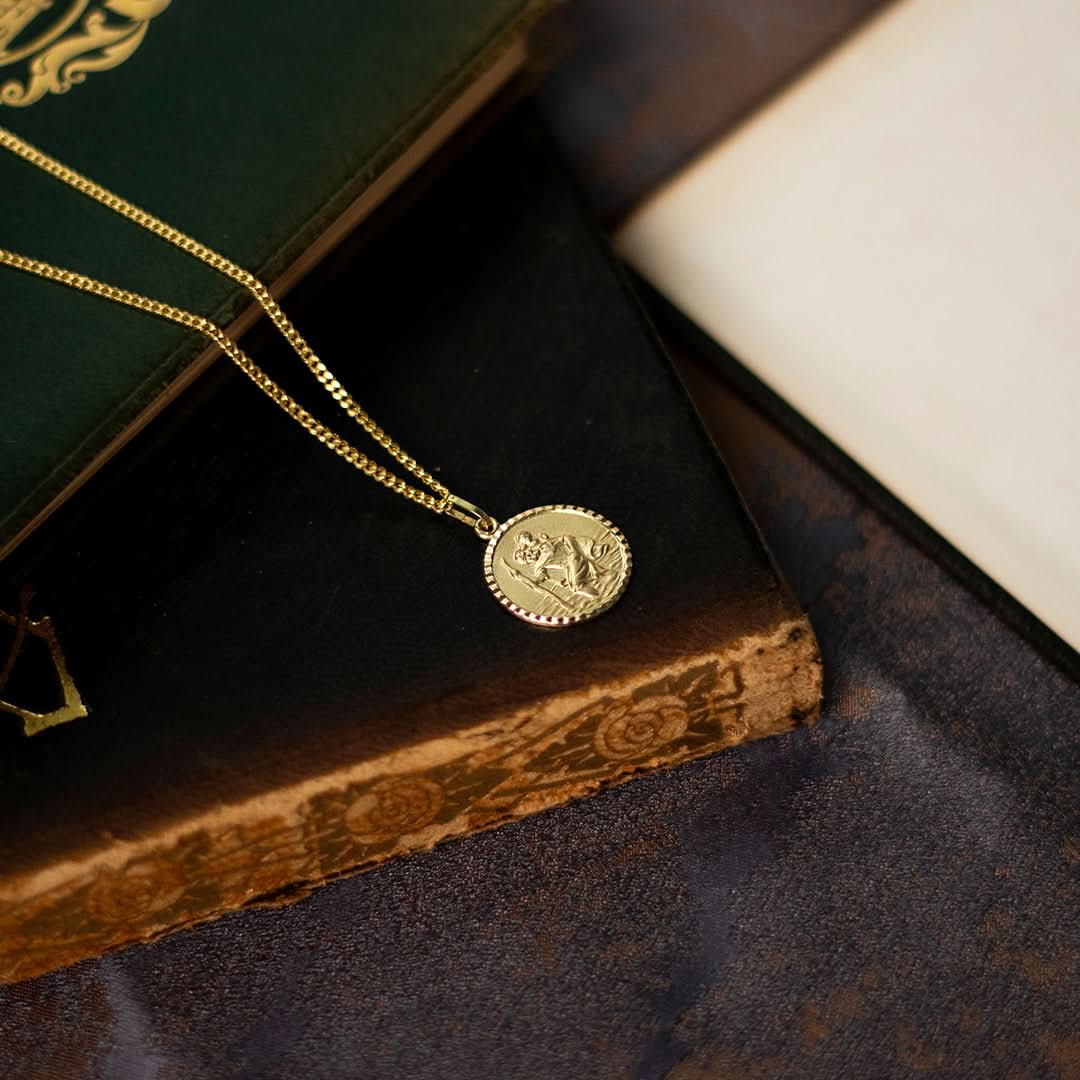 Zoomed-in lifestyle image of a gold St. Christopher necklace. The round pendant with intricate engraving sits naturally on a fine gold chain, lying against a black book