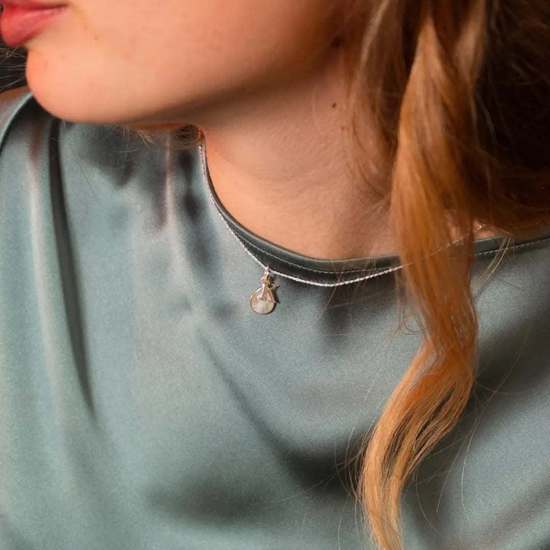 Close-up of a woman wearing a silver necklace with a small moonstone pendant and an initial charm, paired with a teal satin blouse
