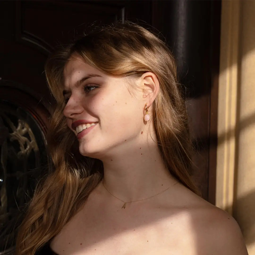 Woman with long hair smiling and wearing gold hoop earrings with pink opal gemstone drops and a gold initial pendant necklace, in natural light