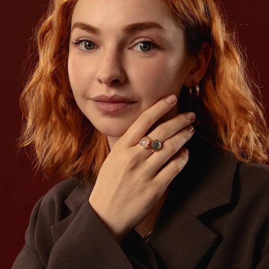 A close-up portrait of a woman with red hair wearing a gold ring with a rose quartz stone and a blue topaz stone, set against a dark red background.