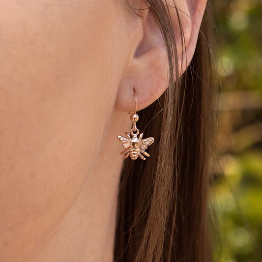 Close-up of a rose gold bee drop earring worn on the ear, featuring a detailed bee charm suspended from a delicate hook fastening.