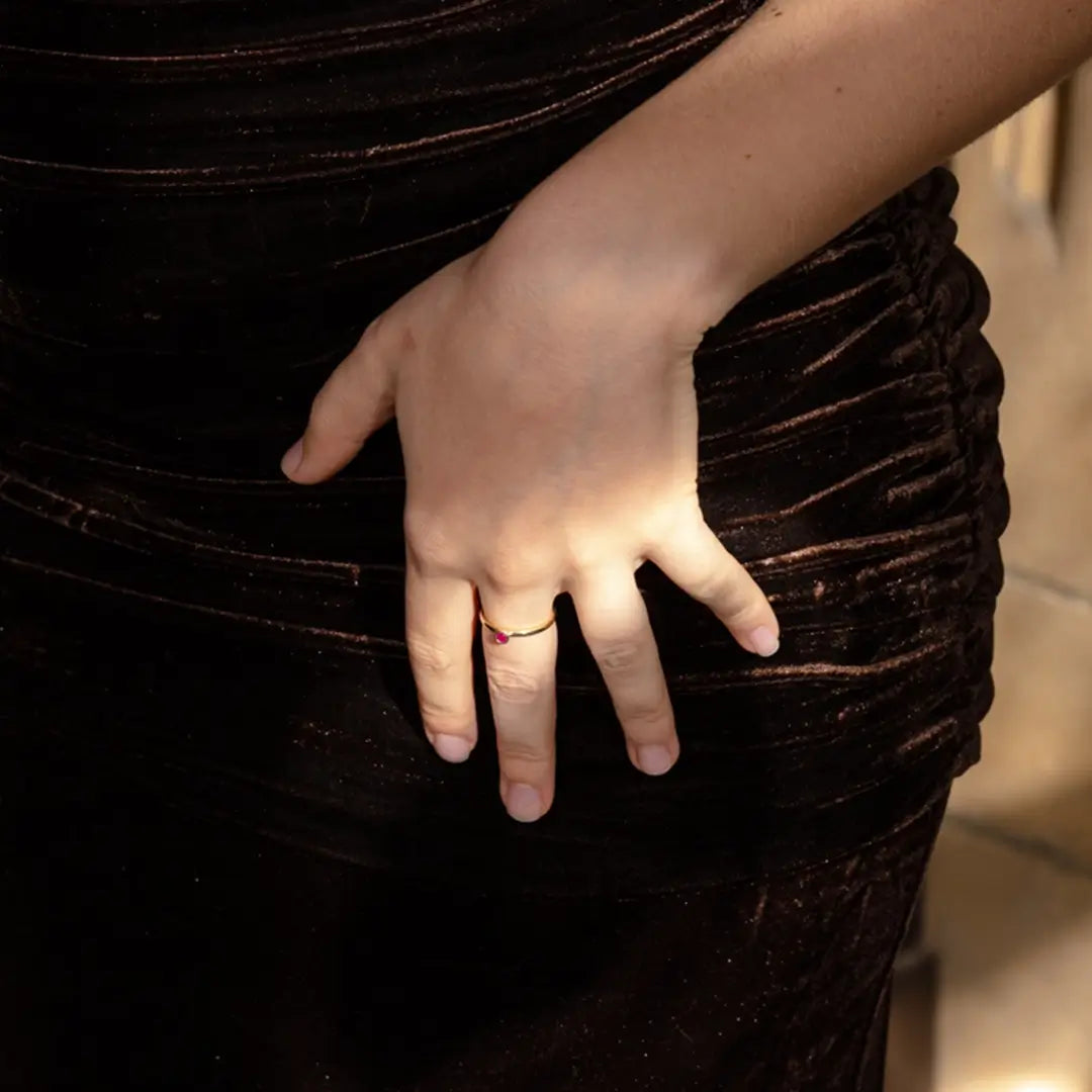 Close-up of a woman’s hand resting on her hip over a ruched dark brown velvet dress. She wears a thin gold ring featuring a small red ruby stone.