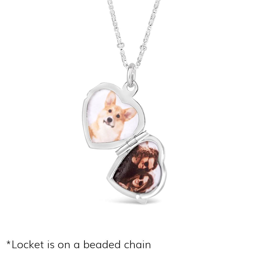 Studio product image of a silver heart-shaped locket necklace, opened to reveal two personalized photos inside: one of a corgi and the other of a couple. The locket is shown on a fine beaded silver chain, photographed against a white background