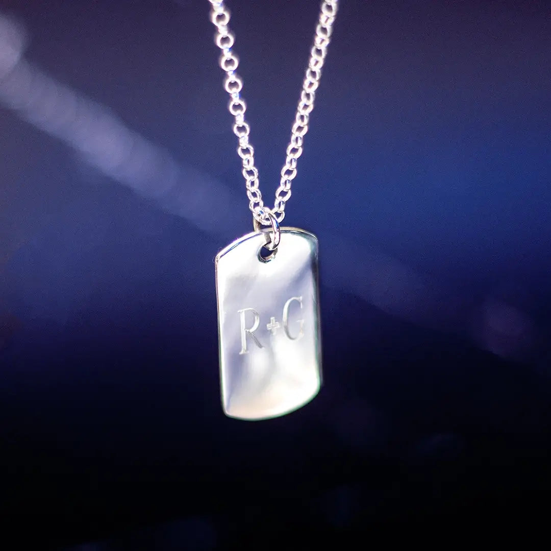 A silver dog tag pendant engraved with the letters “R + G,” hanging from a silver chain against a dark blue gradient background.
