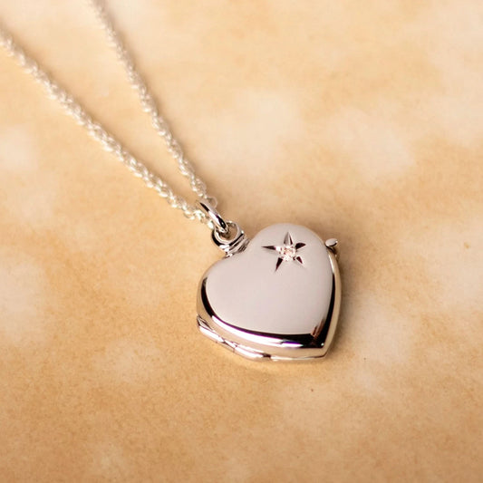lifestyle photo of a sterling silver heart-shaped locket necklace featuring a starburst engraving with a central diamond. Displayed on a rope silver chain and placed on a warm, neutral marbled background