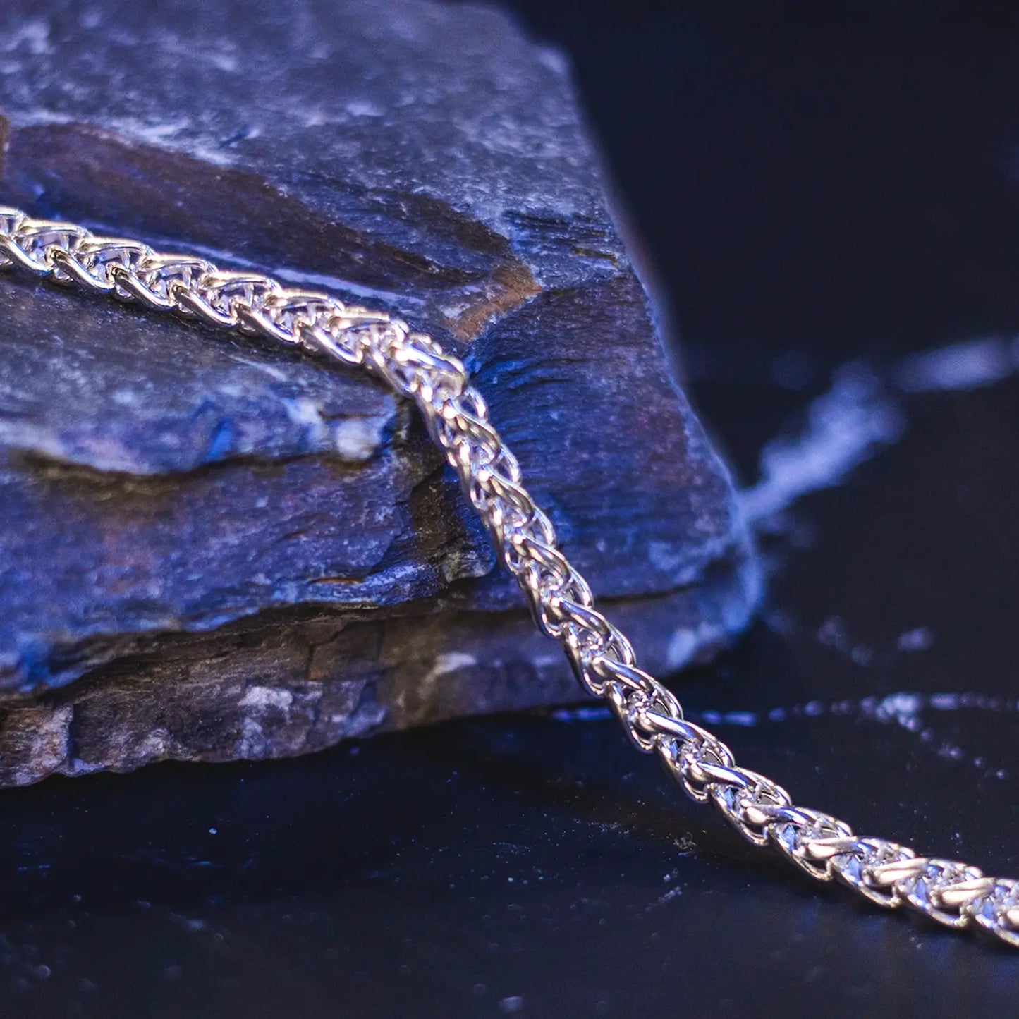 Sterling silver spiga chain displayed on a dark rock surface with reflective blue lighting, highlighting the intricate twisted link design