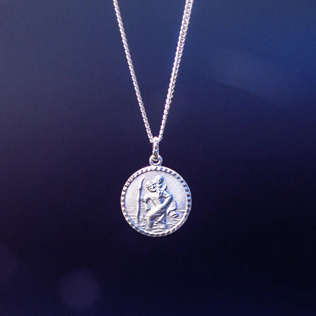 A silver round pendant necklace featuring a raised depiction of Saint Christopher carrying a child, hanging against a dark blue gradient background.