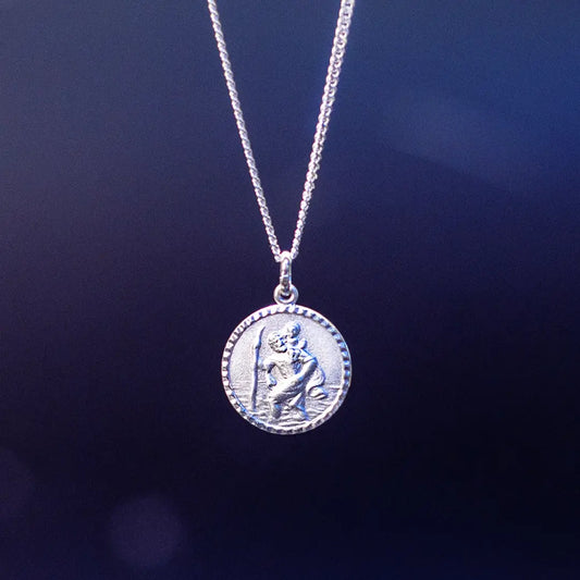 A silver round pendant necklace featuring a raised depiction of Saint Christopher carrying a child, hanging against a dark blue gradient background.