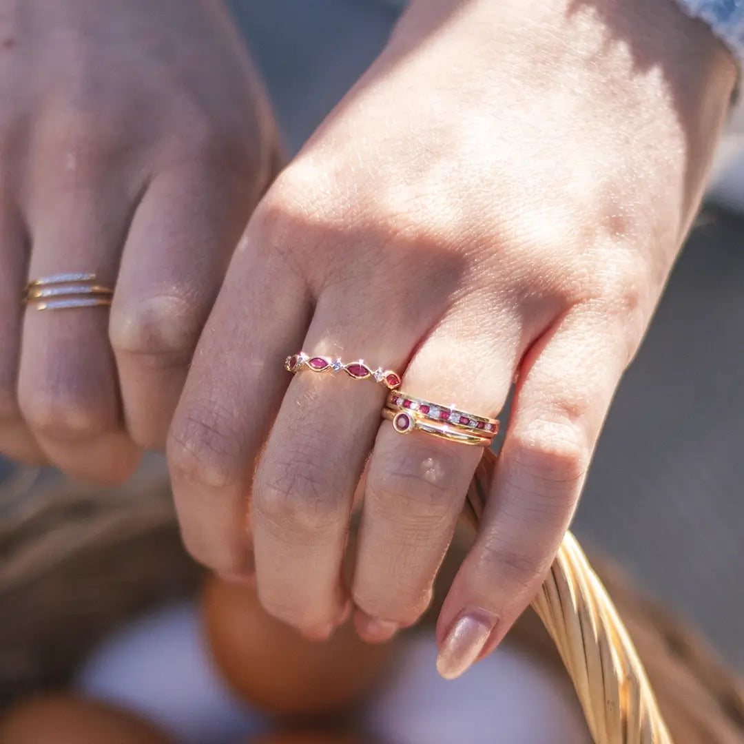 A close-up of two hands wearing gold rings; one hand features multiple gold rings with ruby and diamond details, while the other wears a simple gold band with small diamonds, with a wicker basket in the background.