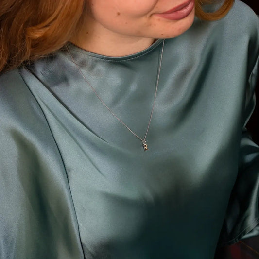 Close-up view of a woman wearing a teal satin blouse and a white gold necklace with a small letter pendant. Her hair falls over her shoulder as she looks slightly down.