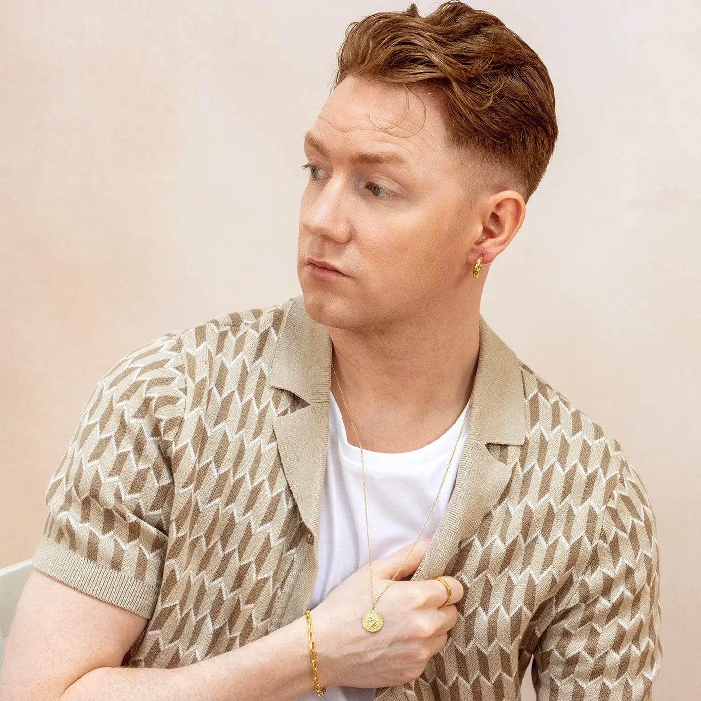 Man dressed a patterned shirt and beige jacket against a plain background, he is also wearing a gold st Christopher necklace