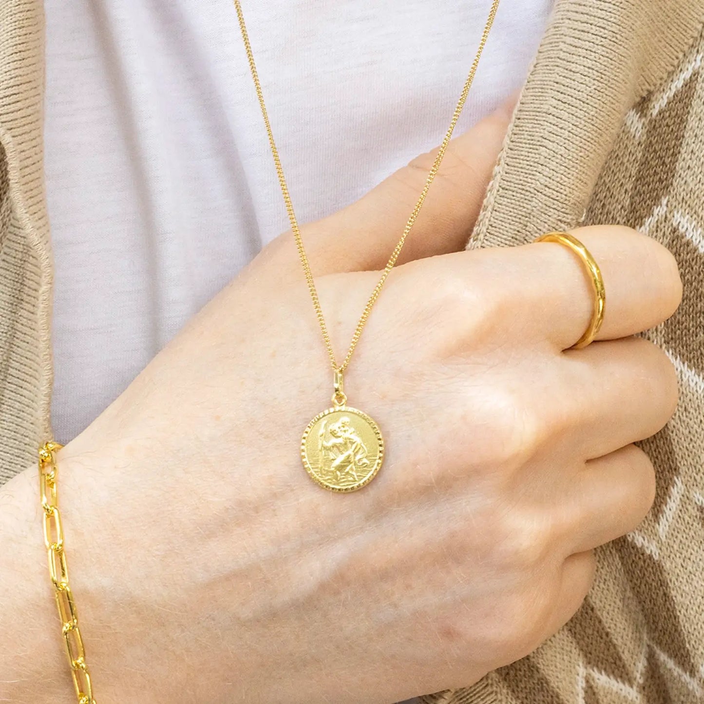 male hand wearing a gold st Christopher necklace with a round pendant, ring, and bracelet against a neutral background.