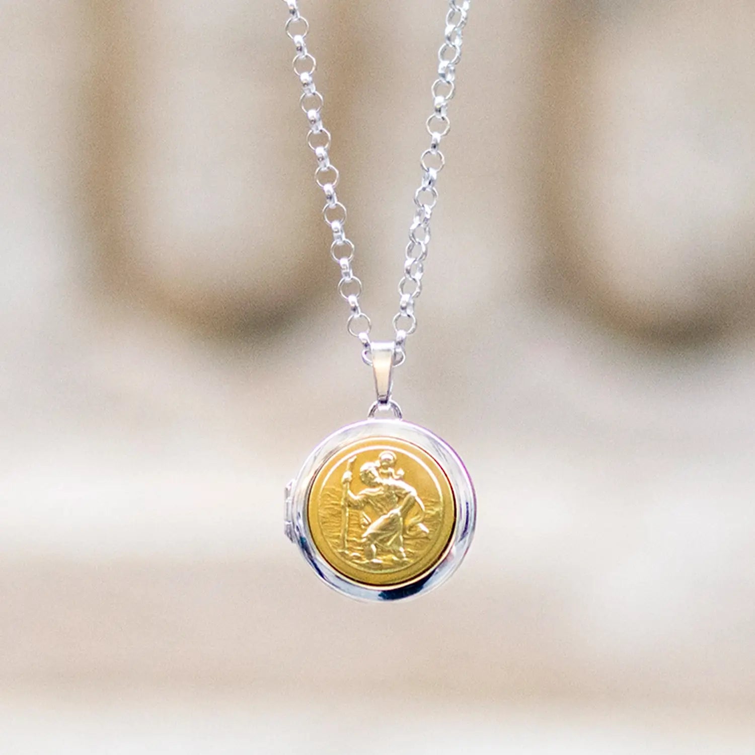 photograph of a silver and gold st Christopher locket from the front hanging on a silver chain against a white stone pillar