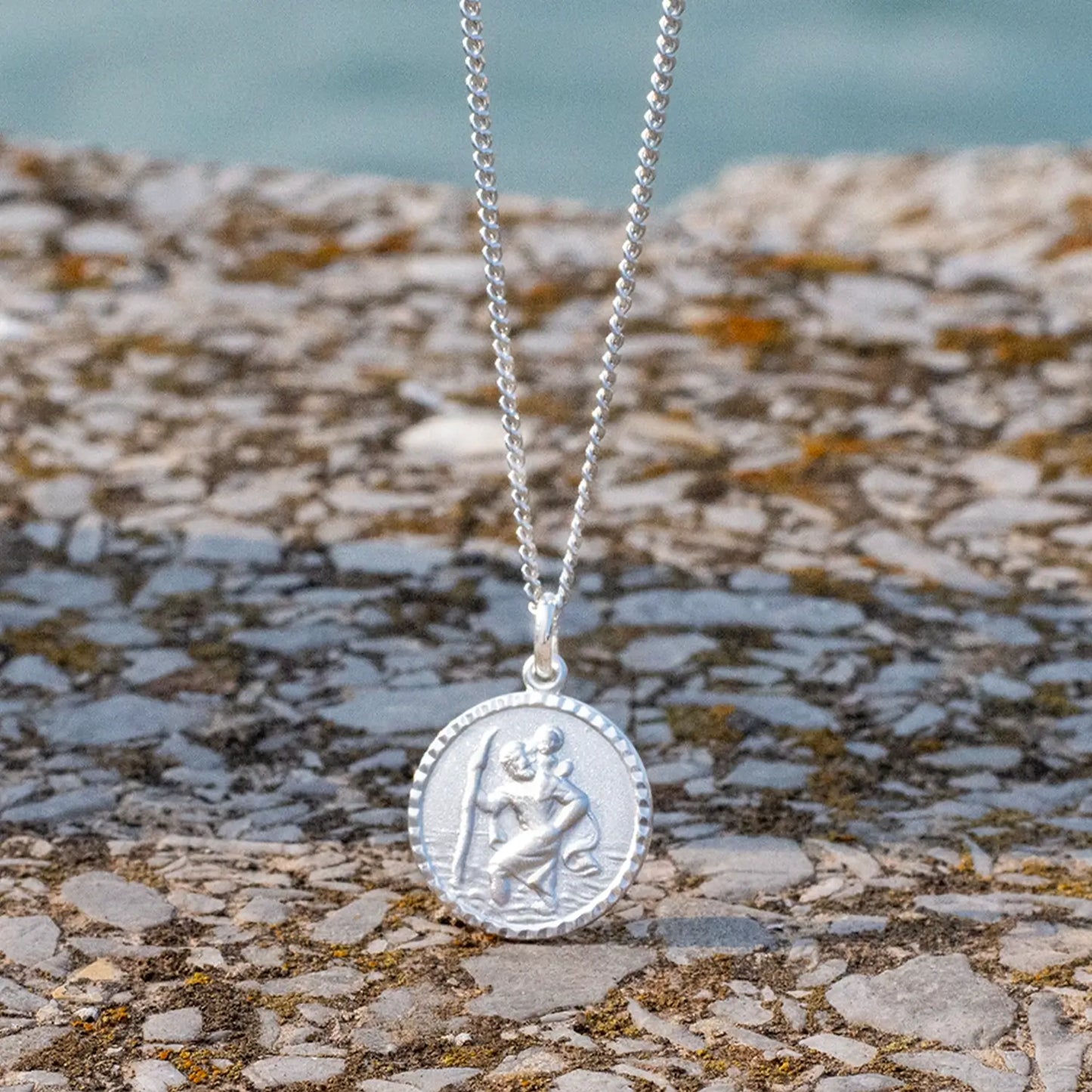 photograph of a silver st Christopher necklace on a curb chain framed against a river and warm stone wall