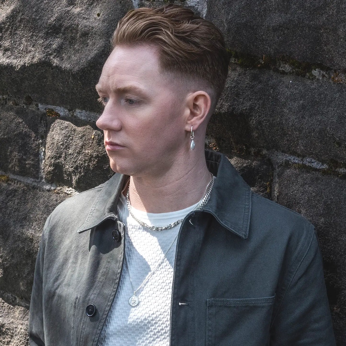 Man wearing a dark jacket and white shirt against a stone wall, wearing a silver st christopher pendant