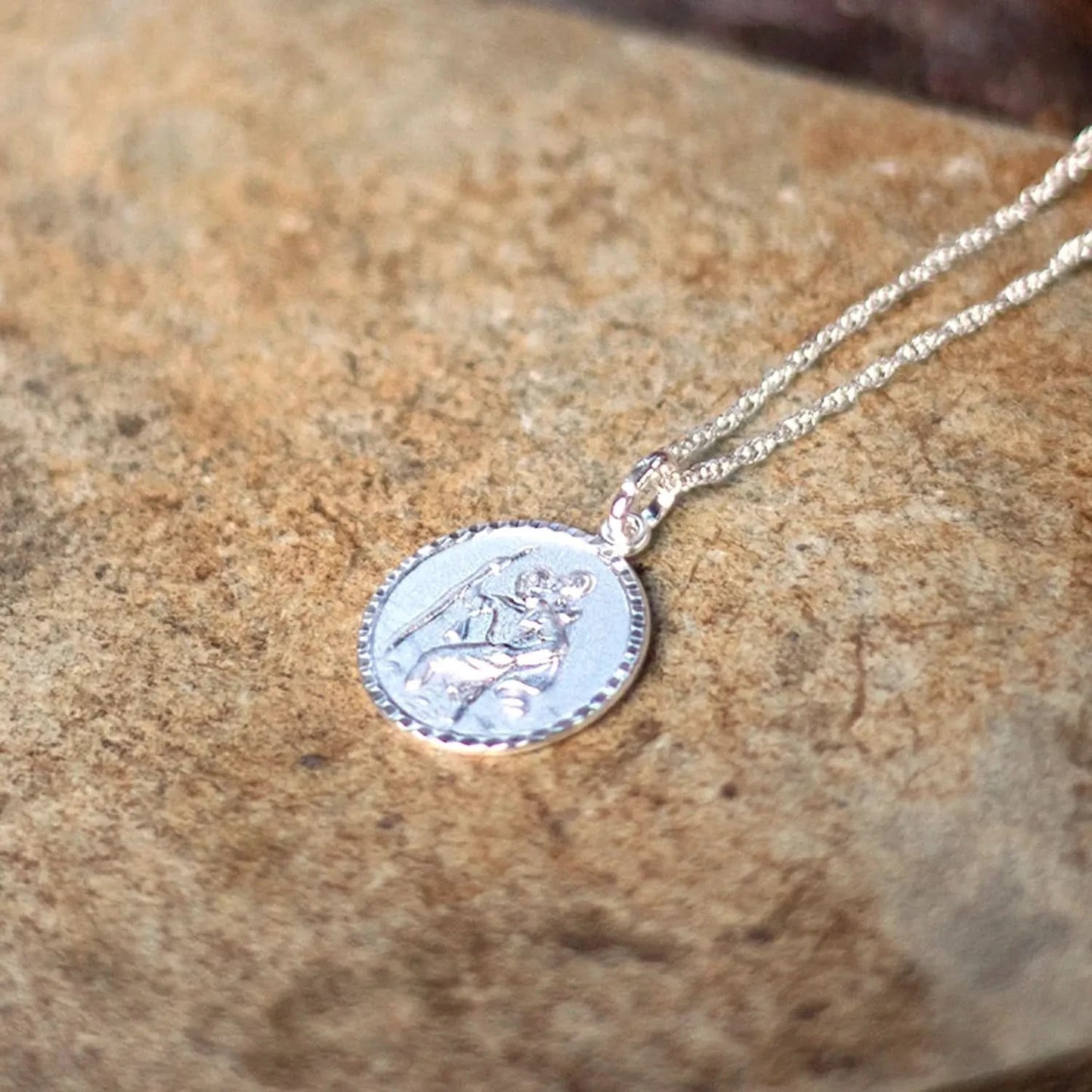 photo of a silver St. Christopher necklace with a circular engraved medallion pendant, displayed on a beige stone. The necklace features a polished silver finish and fine chain