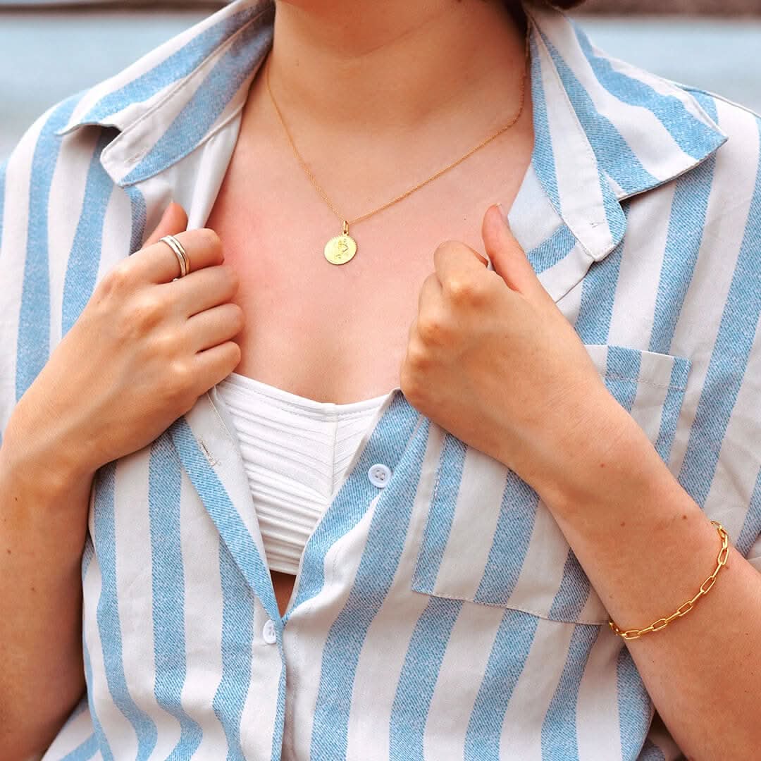 Lifestyle photo of a woman wearing a gold St. Christopher pendant on a fine chain, styled with a blue-and-white striped shirt and gold chain bracelet