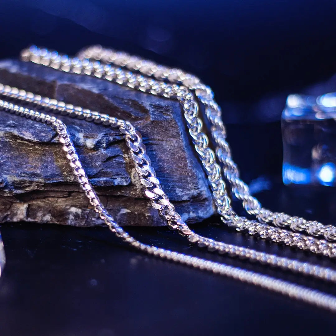 Various sterling silver chain styles, including curb and rope designs, displayed on a dark rock surface with reflective blue highlights