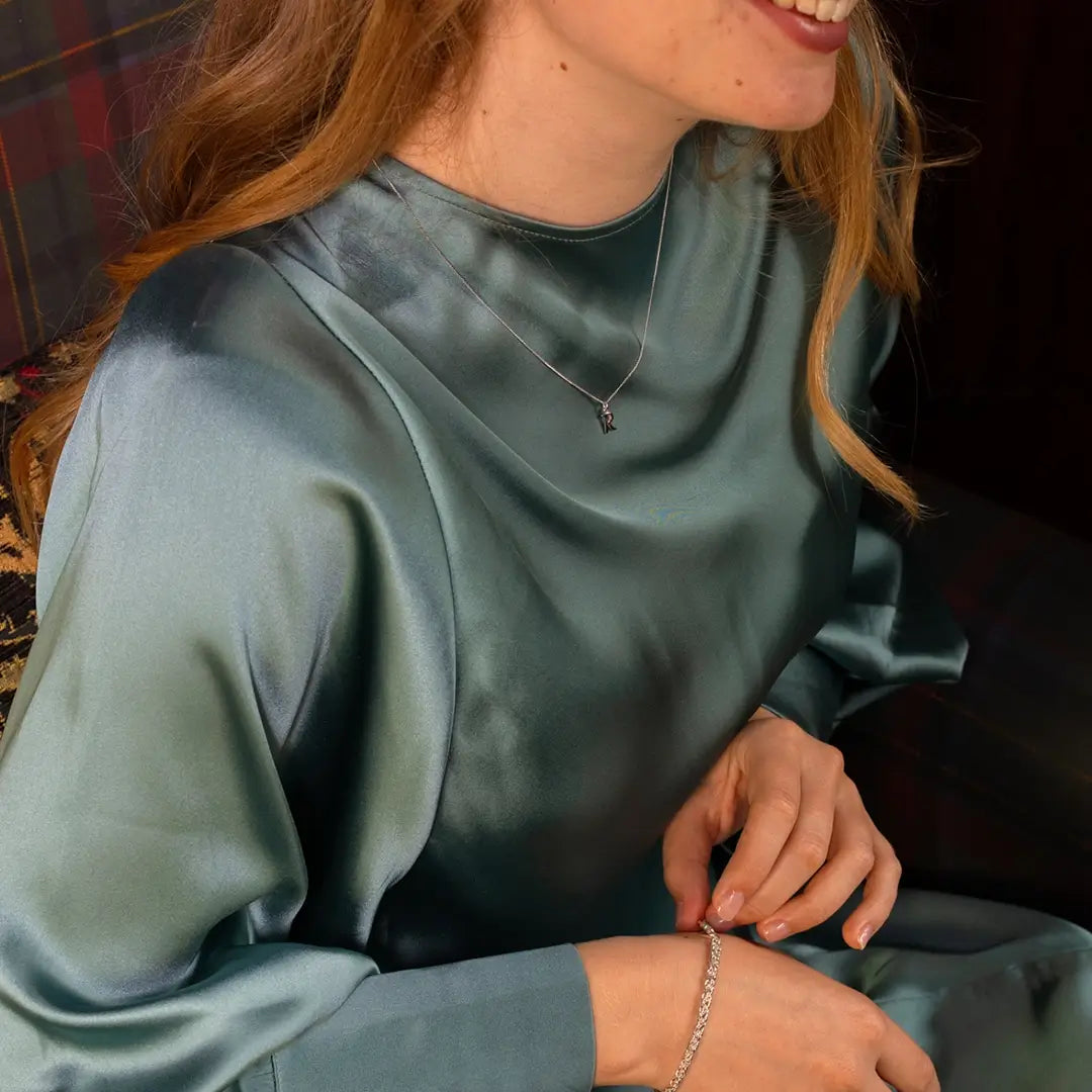Close-up of a woman wearing a light teal satin blouse, sitting on a patterned armchair. She has a delicate white gold initial pendant necklace and a slim bracelet on her wrist.